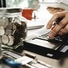 A person uses a calculator while holding a tablet at a desk covered with jars of coins