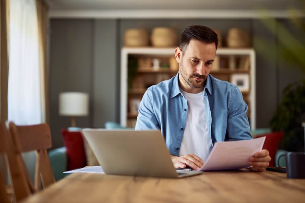A concentrated adult man reading and analyzing a document while sitting in front of a laptop