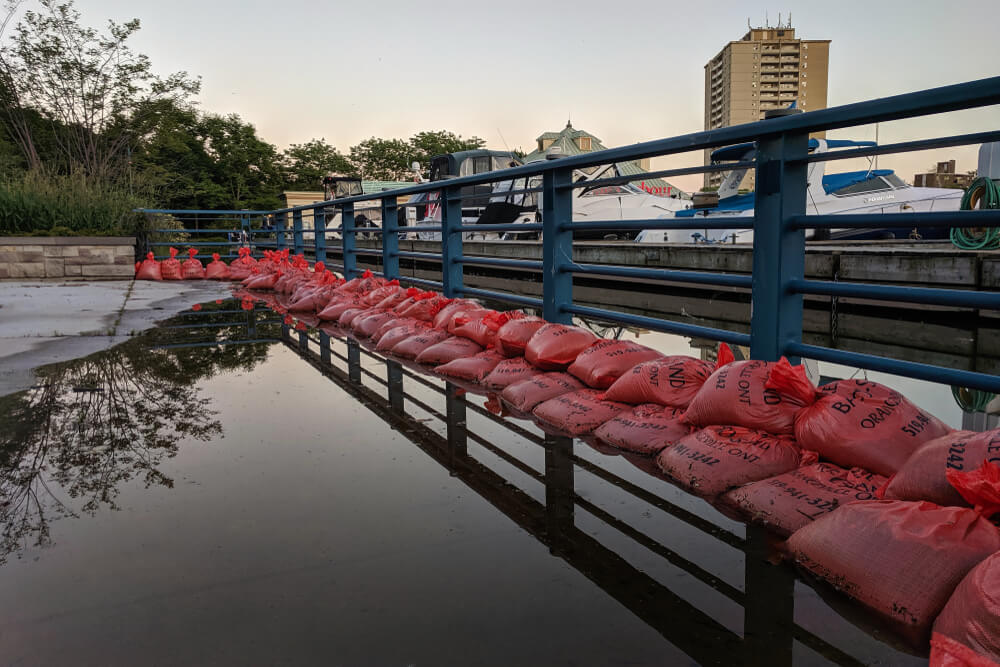 Flooded sidewalk at Port Credit covered with stacked orange sand bags flood barrier as water level rises and overflow in Lake Ontario