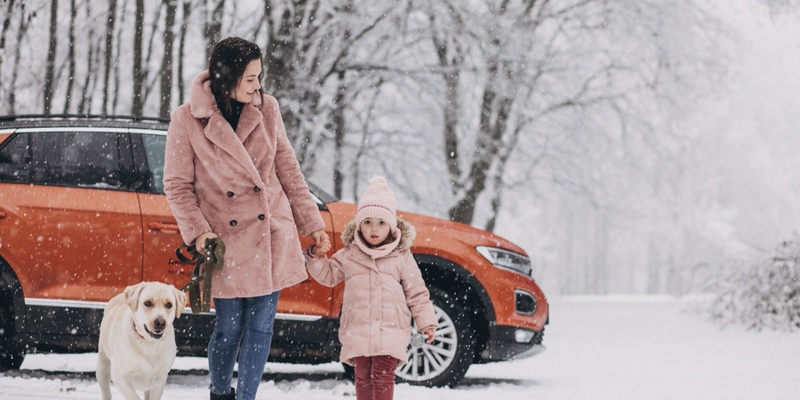 A woman dog and child walk by an orange SUV in the snow
