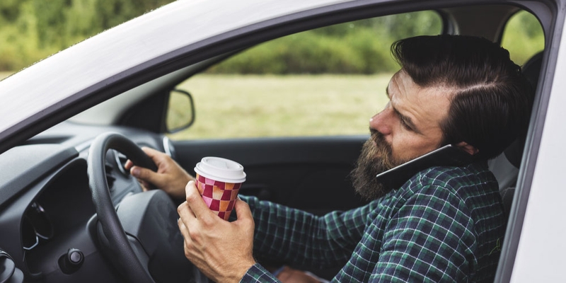 A driver holds a cellphone to hear ear with his shoulder and a coffee cup in one hand and the wheel of his car in the other