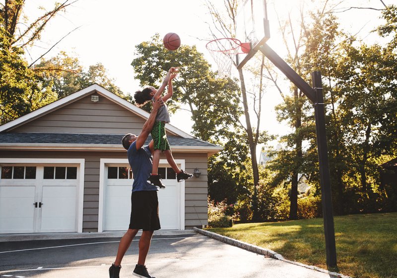 A dad picks up his daughter and holds her up high so she can score a basket at their driveway basketball hoop