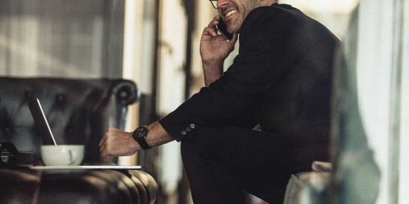 A professional man chats on the phone with a big cappuccino beside him and his laptop not far from reach