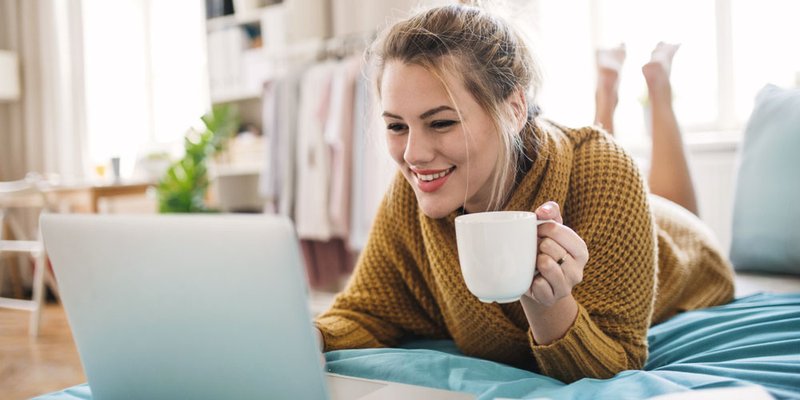 Woman lays on bed casually browsing on her laptop and drinking a coffee