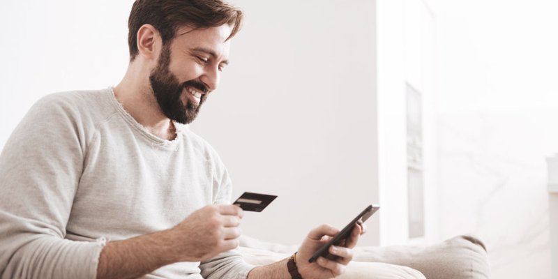 A man sits on a couch and types his credit card details into his phone