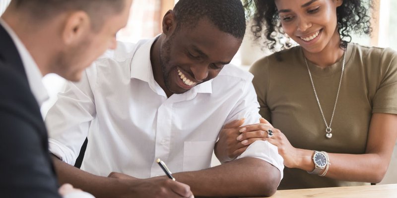 A man signs paperwork while a woman embraces his arm