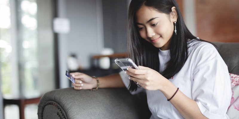 A young woman sits on a couch and shops using her phone and a credit card