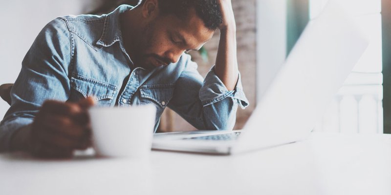A man holds his head in one hand looking overwhelmed as he works on his computer