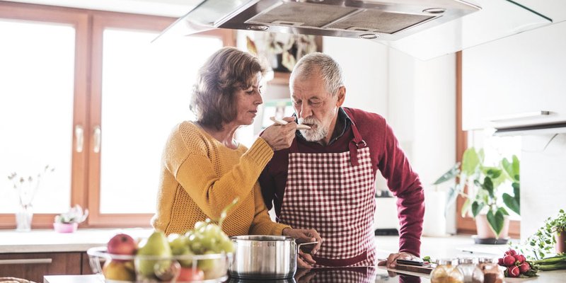 An older woman lets a man taste sauce off a wooden spoon in the kitchen