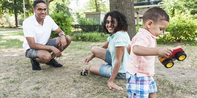 Young family plays at city park and the baby son plays with a toy truck